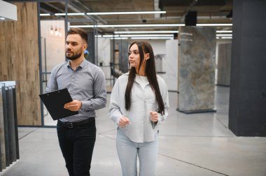 Satış elemanı elinde bir pano tutuyor, büyük bir hırdavat galerisindeki evi için yeni fayanslar yapan bir bayana danışmanlık yapıyor.