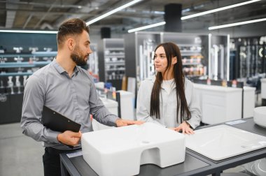 Erkek satış asistanı, modern seramik lavabonun özelliklerini banyo malzemeleri dükkanındaki kadın müşteriye açıklıyor.