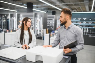 Nalbur dükkanındaki kadın müşterilere banyo lavabosu seçeneklerini gösteren satış asistanı, ev geliştirme ve yenileme seçenekleri hakkında tavsiye veriyor.