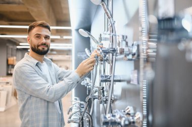 Adam yeni tesisat malzemeleri alıyor, hırdavatçı reyonunda duş başlıklarını kontrol ediyor. Müşteri ev banyosunu yeniliyor.