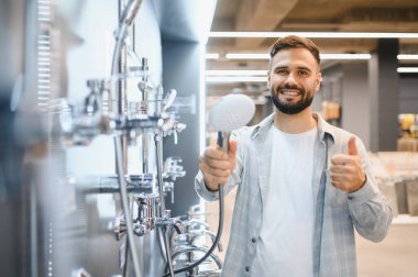 Mutlu adam başparmak kaldırıyor, modern bir tesisat veya banyo mağazasında yeni bir duş sistemi seçiyor.