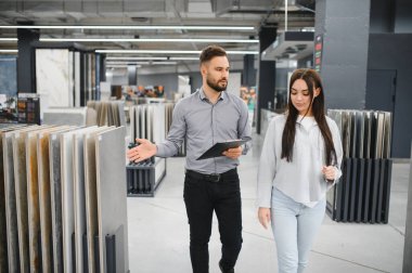 Genç bir bayan müşteriye fayans örnekleri gösteren satıcı, modern bir hırdavatçıda yenileme seçeneklerine yardım ediyor.