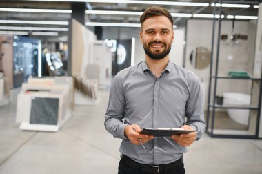 Dost canlısı bir erkek satıcı bir hırdavatçıda elinde bir pano, müşterilere yardım etmek için gülümsüyor.