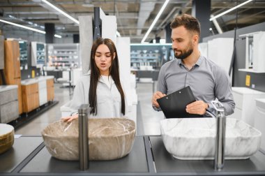 Kadın alıcı, modern bir tesisat veya nalbur dükkanında erkek satış asistanıyla banyo lavabosu seçeneklerini tartışıyor.