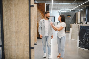 Bir sergi salonunda fayans örneklerini karşılaştıran, döşeme seçeneklerini tartışan ve birlikte bir ev yenileme planı yapan, gülümseyen ve düşünceli bir çift.