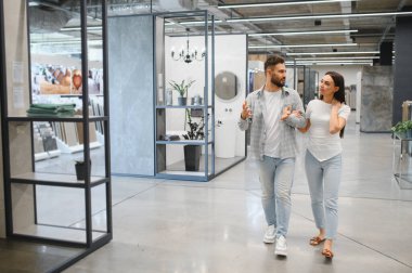 Genç bir çift ev geliştirme galerisinde yürüyor, yenileme ve iç tasarım projeleri seçeneklerini tartışıyorlar.