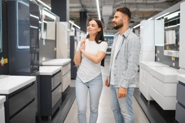 Genç çift alışveriş yapıyor, banyo mobilyalarına bakıyor ve modern bir galeride ev dekorasyonu seçenekleri sunuyor.