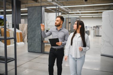 Satıcı ve kadın bir galeride ev yenileme projesini tartışıyorlar, zemin ve duvar örtülerini seçiyorlar.