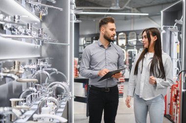 Erkek satış asistanı elinde bir pano, hırdavatçıdaki kadın müşteriye tesisat tesisatında yardım ediyor. Yeni banyo ya da mutfak ekipmanları almak