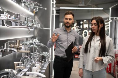 Satıcı, genç bir bayana modern bir tesisat bölümünde banyosu ya da mutfağı için musluk seçmesinde yardım ediyor.