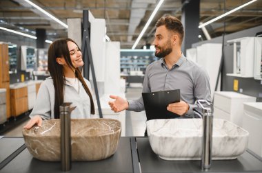 Kadın müşteri ve erkek satıcı, modern bir hırdavatçı dükkanında banyo demirbaşlarını tartışıyor.