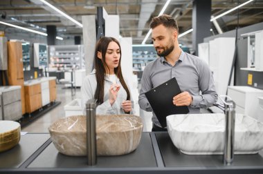 Müşteri ve satıcı bir hırdavatçıda banyo yenileme seçeneklerini tartışıyor, farklı lavabo modellerine odaklanıyor