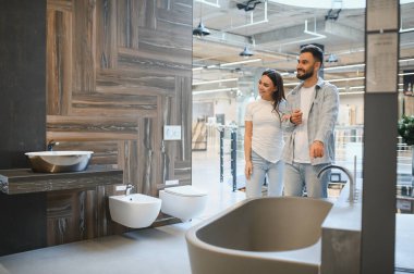 Genç, gülümseyen bir çift modern hijyen malzemelerini, fayanslarını ve mobilyalarını seçiyorlar. Bir banyo galerisinde ev geliştirme projeleri için.