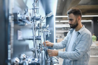 Genç adam modern bir hırdavatçı dükkanından banyo malzemeleri alıyor, duş başlıkları ve muslukları inceliyor.