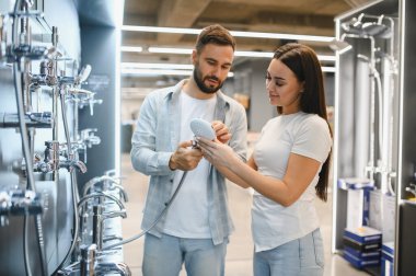 Genç çift, bir tesisat dükkanında duş başlıklarını ve banyo demirbaşlarını karşılaştırıyor ve evlerinin tadilatı için karar veriyorlar.