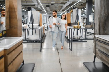 Genç çift kol kola yürür, banyo mobilyalarını ve lavaboları seçer, ev dekorasyonu için plan yapar.