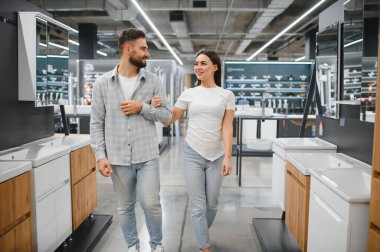 Genç çift kol kola yürüyor, banyo süslemelerine bakıyor ve modern bir ev geliştirme galerisinde batıyor.