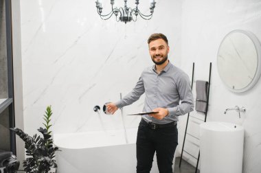 Parlak galeride gülümseyen satıcı elinde tablet, modern beyaz küveti sunuyor ve temiz, zarif bir banyo vitrininde batıyor.