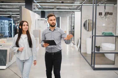 Elinde bir pano tutan satıcı, modern bir mağazada kadın bir müşteriye ev geliştirme ürünlerini seçmesinde rehberlik ve yardım ediyor.