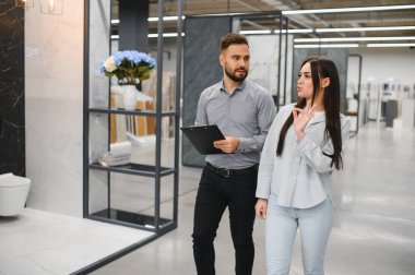 Modern bir sergi salonunda satış asistanına danışmanlık yapan bir kadın, banyo veya ev yenileme malzemeleri seçiyor.