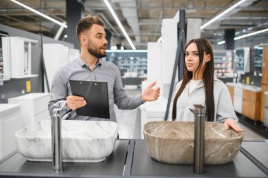 Hırdavatçıda yeni lavabo seçen kadın müşterilere yardım eden satıcı, ev yenileme fikirlerini tartışıyor.