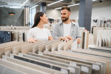 Parlak bir sergi salonunda genç bir çift evlerini yenilemek için döşeme örnekleri seçiyor, gülümsüyor ve yeni iç mekanlarını planlıyorlar.