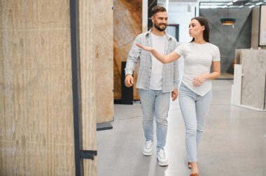 Fayans dükkanında yürüyen ve konuşan genç bir çift, ev yenileme projeleri için malzeme seçiyorlar.