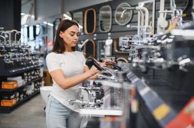 Hırdavatçıda duran bir kadın, banyo veya mutfak yenileme projesi için farklı muslukları dikkatle inceliyor.