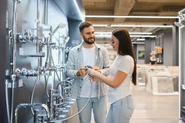 Duşlara ve musluklara bakan genç bir çift, hırdavatçıda banyo tadilatı için tesisat malzemeleri seçiyorlar.