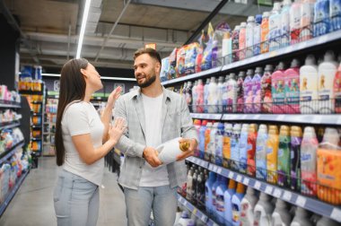 Genç çift, marketin çamaşır deterjanı koridorunda ev ürünleri hakkında hararetli tartışmalar yapıyor.