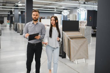 Satıcı, modern bir perakende galerisinde ev yenileme projesi için yeni fayanslar seçen bir kadına yardım ediyor.