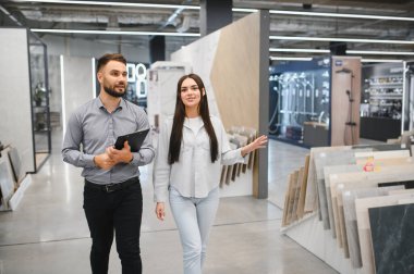 Modern bir perakende mağazasında döşeme ve duvar döşemesi seçen genç bir kadına yardımcı olan erkek satış asistanı.