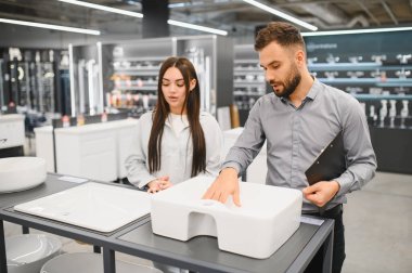 Satış elemanı, kadın müşterinin farklı lavabolarını bir hırdavatçıda ya da ev geliştirme merkezinde gösteriyor.