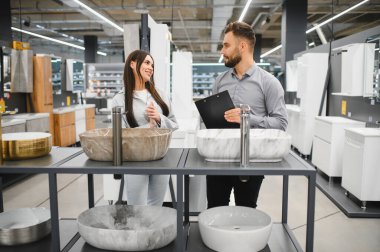 Kadın müşteri ve erkek satıcı bir hırdavatçı dükkanında banyo lavabolarını tartışıyor, ev geliştirme stillerine odaklanıyor.