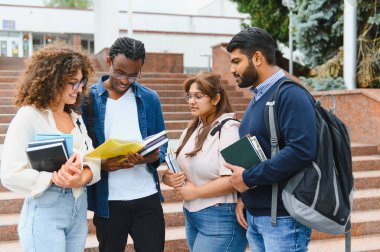 Group of diverse university students standing on campus stairs, discussing study materials and reading books together