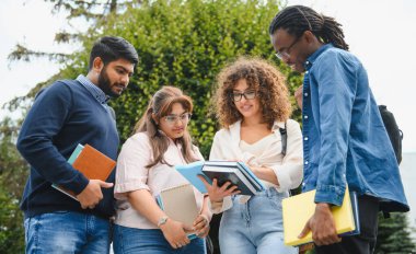 Group of diverse college friends collaborating on an assignment outdoors, holding books and discussing school work