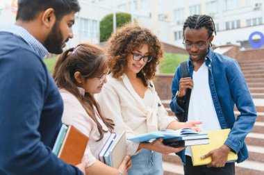 Four young diverse university students standing on campus stairs, discussing study materials and sharing books