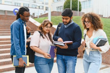Group of multi ethnic students standing outdoors on university campus, reviewing textbooks and discussing lessons