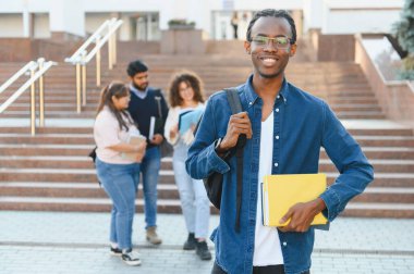 Sırtında kitap ve sırt çantası olan gülümseyen genç bir öğrenci üniversite kampüsünde çeşitli öğrencilerin önünde duruyor.