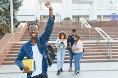 Başarılı ve eğitimi kutlayan neşeli Afrikalı Amerikalı öğrenci, üniversite kampüsünde çeşitli arkadaşlarıyla kitap tutuyor.