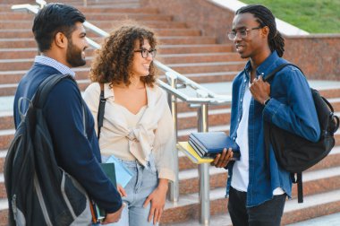 Çeşitli öğrenciler üniversite kampüsünün merdivenlerinde bir araya gelerek tartışıyor ve gülümsüyor, eğitimi, arkadaşlığı ve çeşitliliği temsil ediyorlar.