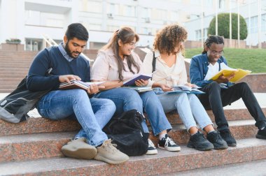 Çok ırklı bir grup genç üniversite öğrencisi kitap paylaşıyor, kampüs basamaklarında öğreniyor, sınavlara hazırlanıyor.