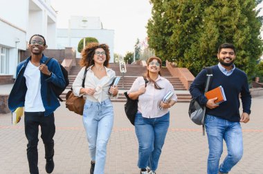 Üniversite kampüsünde kitap ve sırt çantaları taşıyan bir grup mutlu genç öğrenci. Eğitim ve dostluk kavramı gösteriliyor