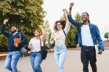 Kitapları ve sırt çantaları olan, yumruklarını kaldıran, üniversite kampüsündeki eğitim başarısını kutlayan bir grup mutlu çok ırklı öğrenci.