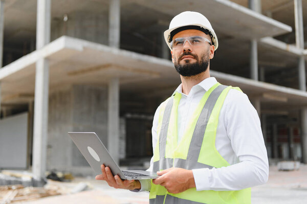 Engineer wearing hard hat and safety vest holding laptop, supervising modern building construction project