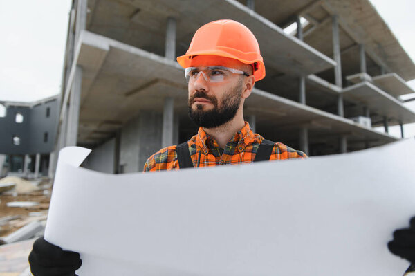 Строитель в каске и защитных очках, держит чертеж на стройке