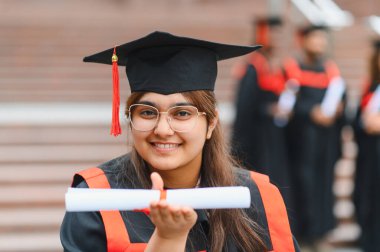 Genç Hintli kadın başarısını kutluyor, mezuniyet elbisesi ve şapkası giyiyor, diplomasını mutlulukla tutuyor.