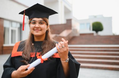 Mezuniyet kepi ve cübbesi takmış Hintli bir kadın. Elinde sarılı diplomasıyla gülümsüyor. Eğitimsel başarısını ve geleceğini kutluyor.