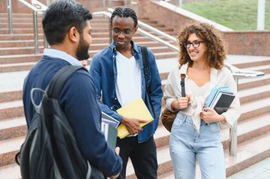 Sırt çantaları ve kitaplar taşıyan, kampüs merdivenlerinde duran ve konuşan çeşitli genç üniversite öğrencileri.