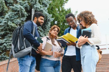 Kampüste duran çeşitli üniversite öğrencileri, kitaplarla ve defterlerle akademik materyalleri tartışıyor ve işbirliği yapıyorlar.